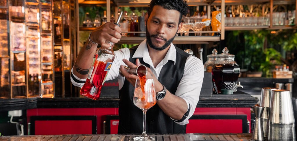 Bartender in a vest pouring Best Happy Hours red liquid into a glass with ice at a bar counter.