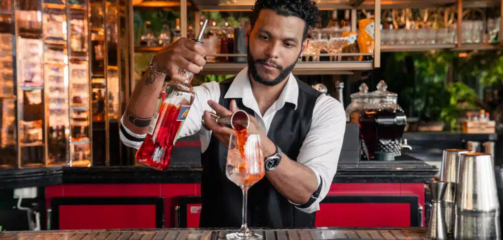 Bartender in a vest pouring Best Happy Hours red liquid into a glass with ice at a bar counter.