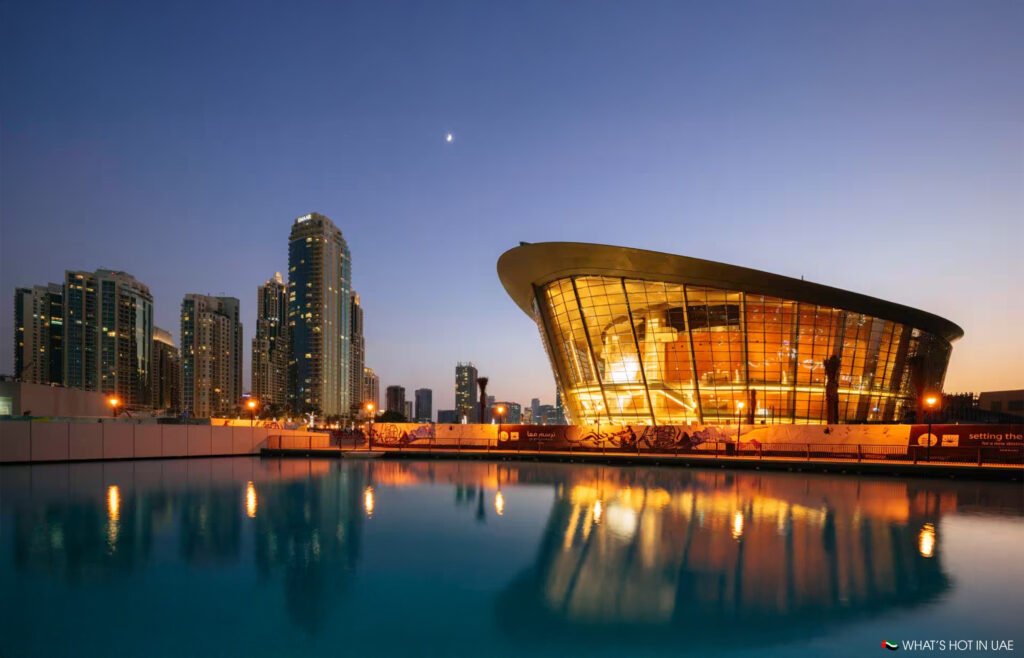 The illuminated Dubai Opera building is reflected in a tranquil water pool at dusk, with city skyscrapers in the background.