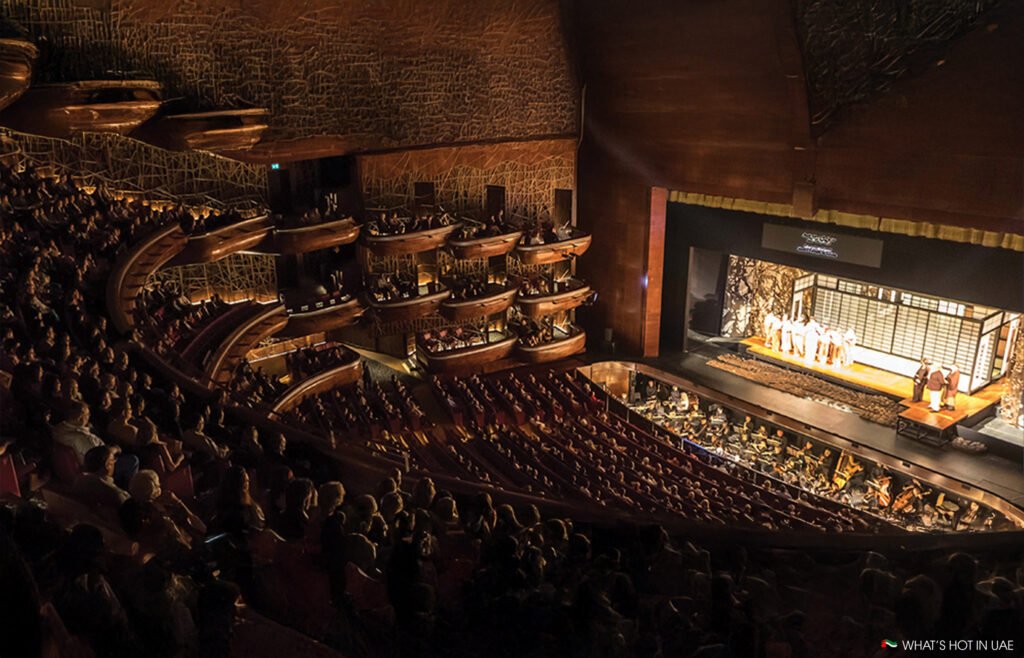 A grand theater with a large audience watching a live stage performance; orchestral musicians are seated below the stage.