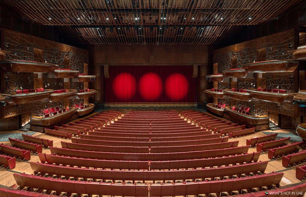 An empty theater with red velvet seats, ornate wooden box seats, and a red curtain on the stage.