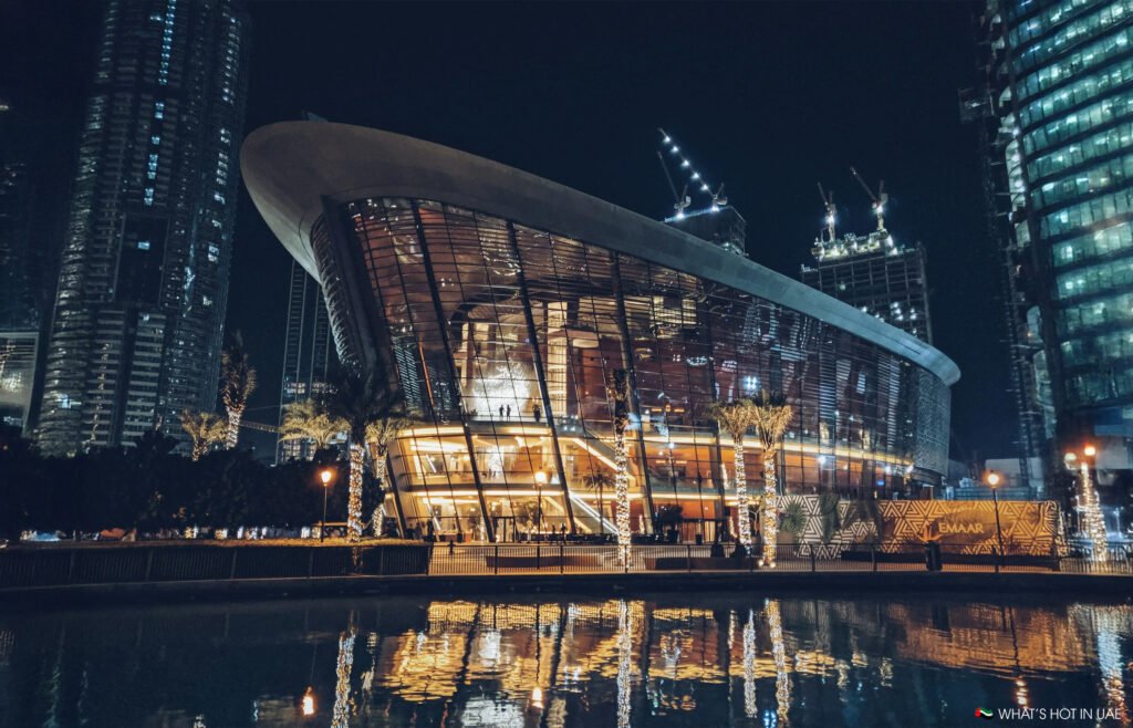 A modern, illuminated building with a reflective glass exterior, surrounded by city skyscrapers at night.