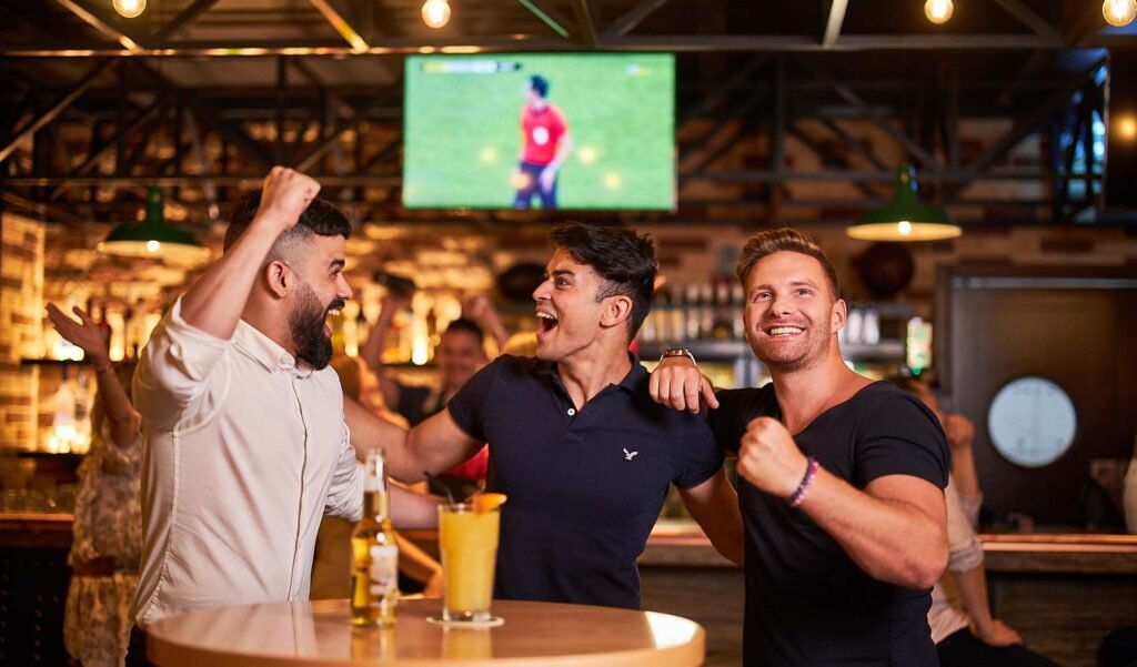 Three men celebrate enthusiastically in a bar with a sports game on a TV in the background.