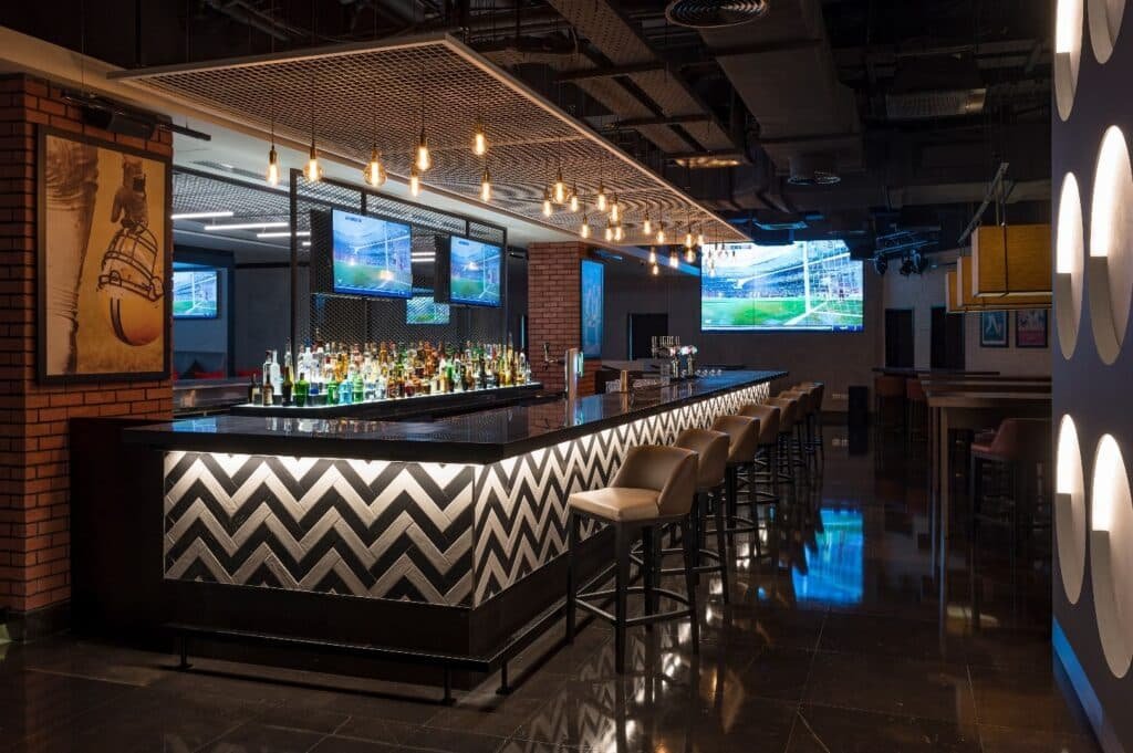 A modern bar with a zigzag-patterned counter, high chairs, and large screens displaying a sports match.