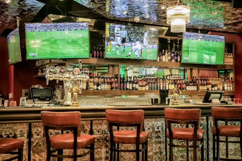 Dubai's Best Sports Bars: Three bar stools in front of a counter with multiple taps, backed by shelves of bottles and three TVs showing soccer games.