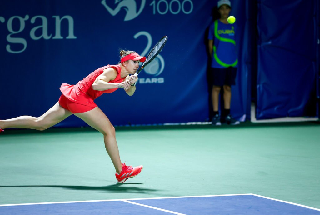 A tennis player in a red outfit lunges to hit a ball during a match, with a ball boy in the background.
