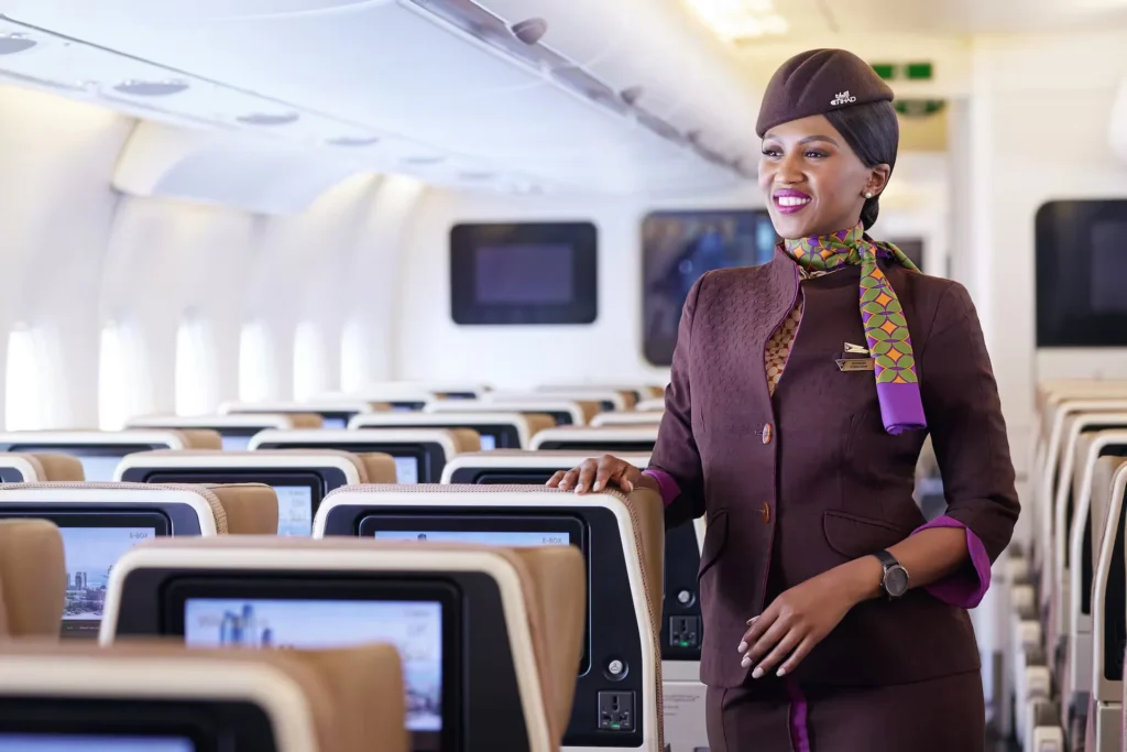 Winner of World's Safest Airlines 2026, Etihad Airways cabin crew member smiling inside an aircraft cabin, standing beside rows of economy seats with seatback screens.