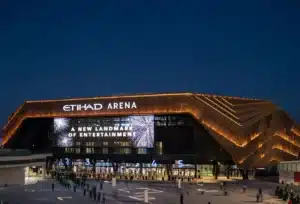 Etihad Arena illuminated at night with prominent digital signage and people gathered at the entrance.