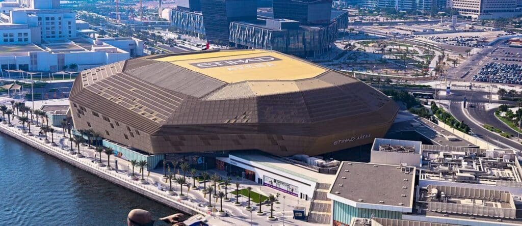 Aerial view of Etihad Arena, a large octagonal building by the waterfront, surrounded by modern cityscape.
