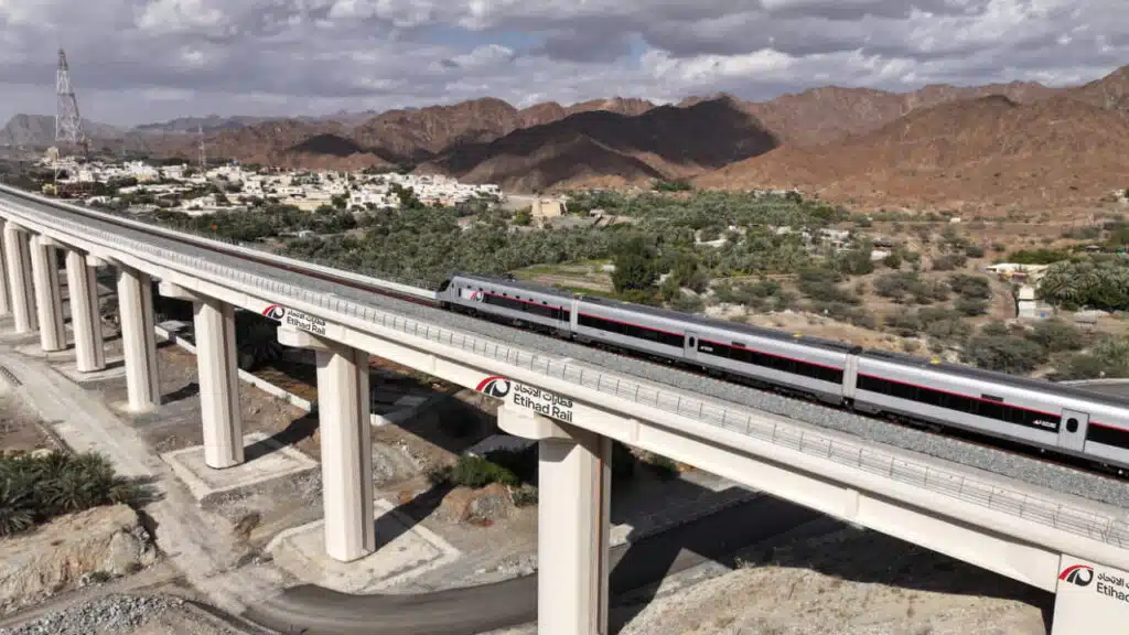 A train travels on an elevated rail bridge with mountains and a small town in the background.