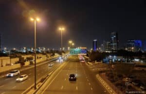 A night view of a busy highway in an urban area, with illuminated streetlights and skyscrapers in the background.