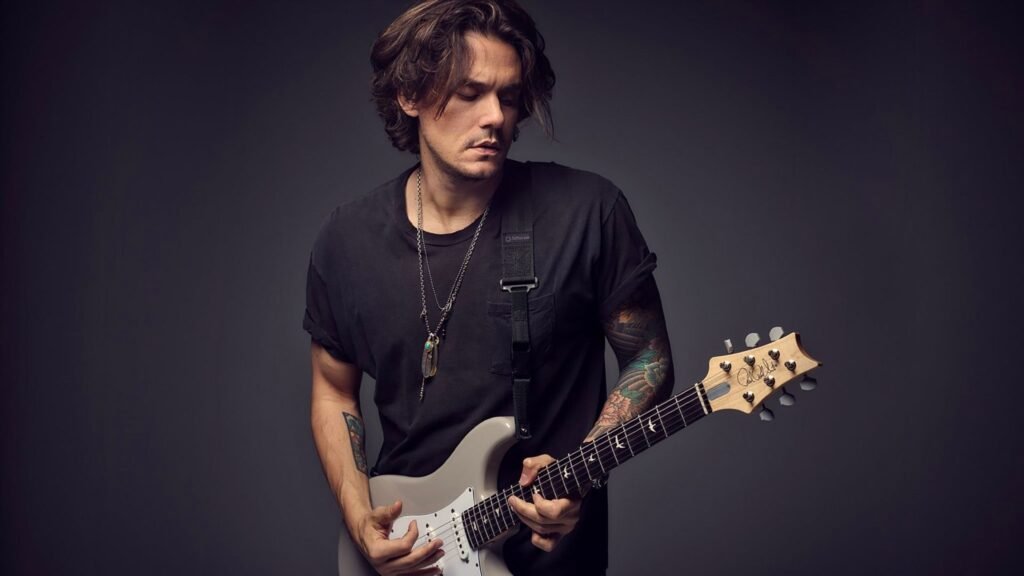 John Mayer playing a guitar in front of a gray background.