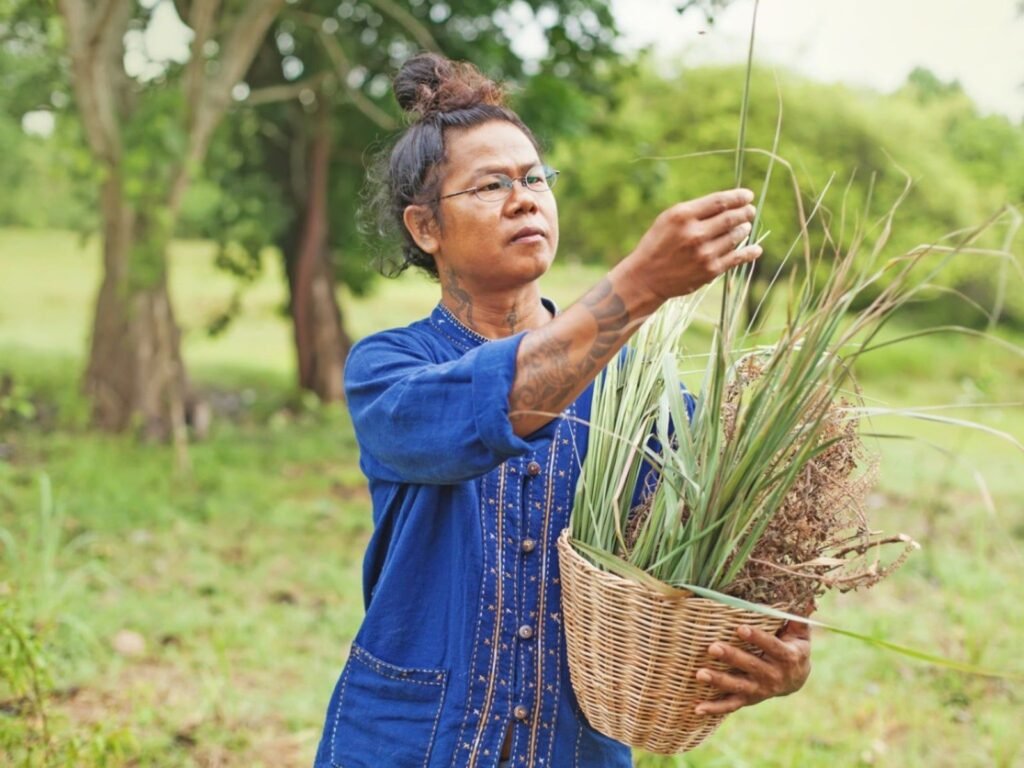 A person in a blue embroidered shirt holds a basket of grasses in a lush, green outdoor setting.