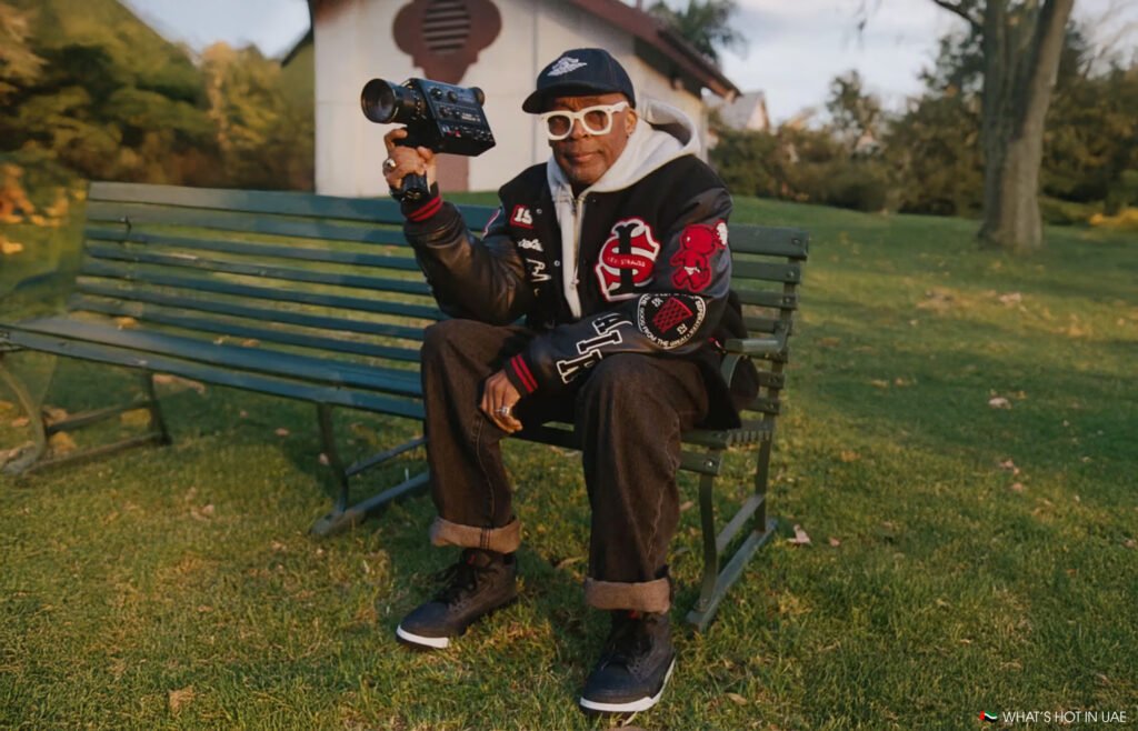 Spike Lee sitting on a park bench holding a camera, wearing a black jacket with red and white patches, white glasses, and a cap.