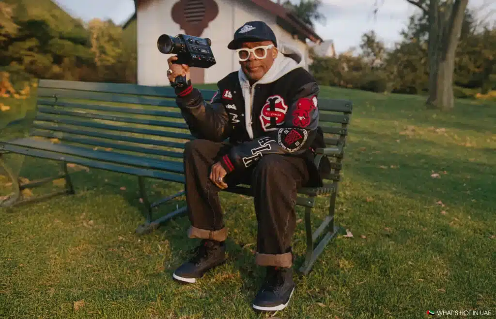 Spike Lee sitting on a park bench holding a camera, wearing a black jacket with red and white patches, white glasses, and a cap.