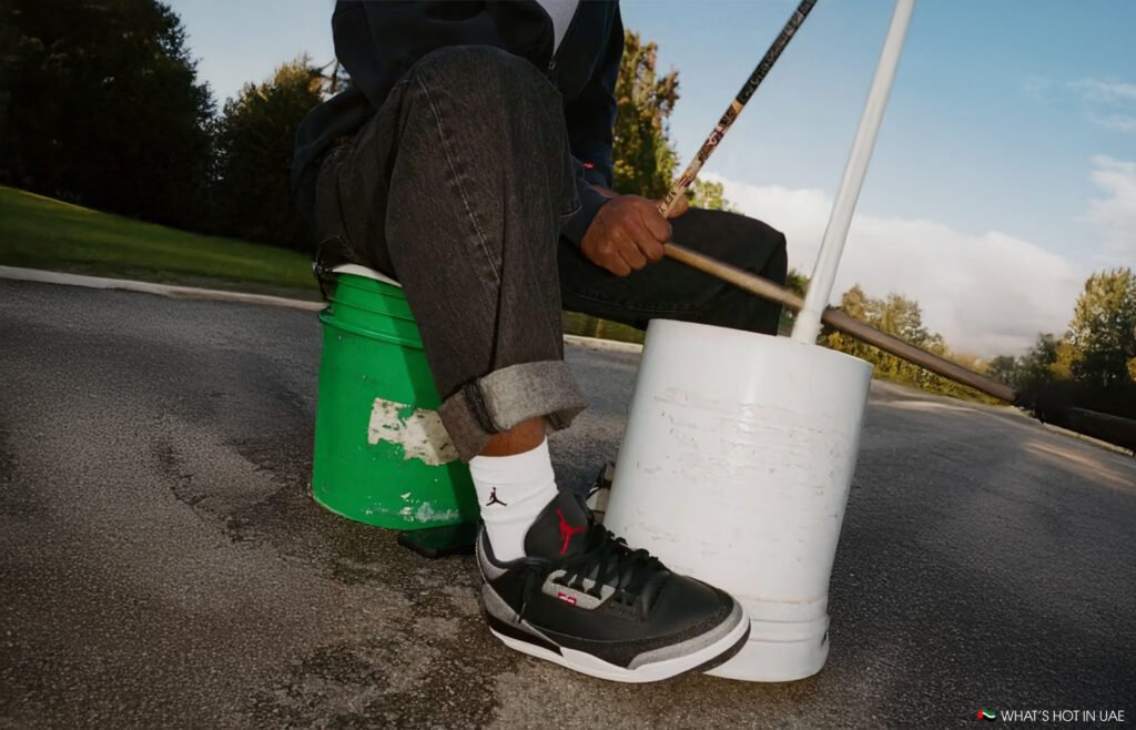 Person playing drums on buckets, wearing rolled-up jeans and sneakers, sitting on a green and white bucket.