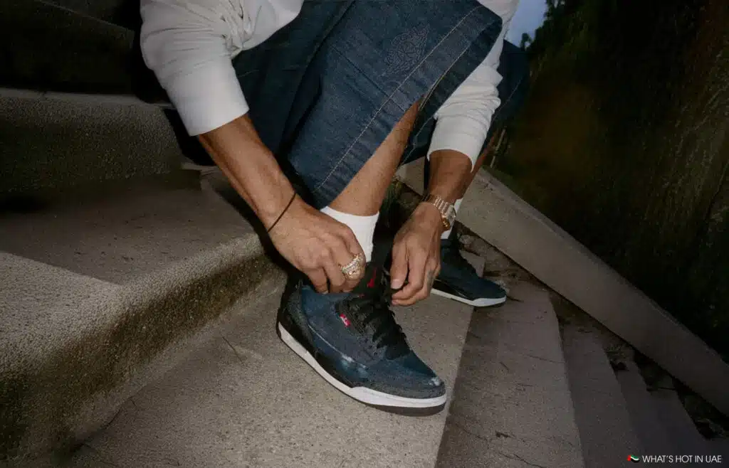A person sitting on steps laces up a Levi's x Jordan Brand blue sneaker, wearing an off-white sweater and denim.
