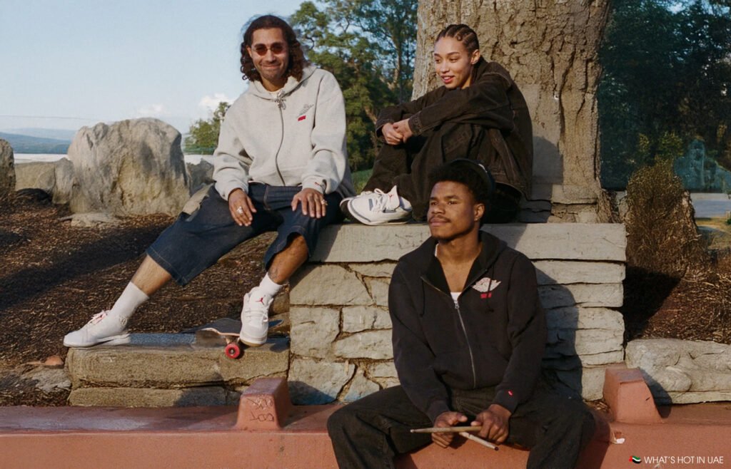 Three people wearing casual clothing sit together outdoors, one holding a skateboard, another with drumsticks.