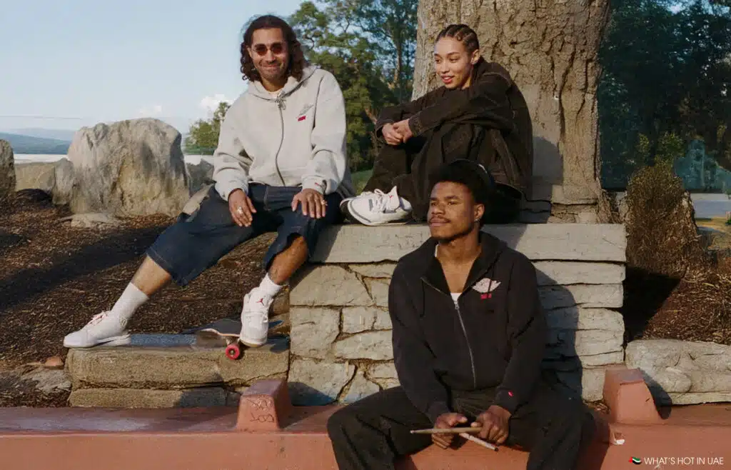 Three people wearing casual clothing sit together outdoors, one holding a skateboard, another with drumsticks.