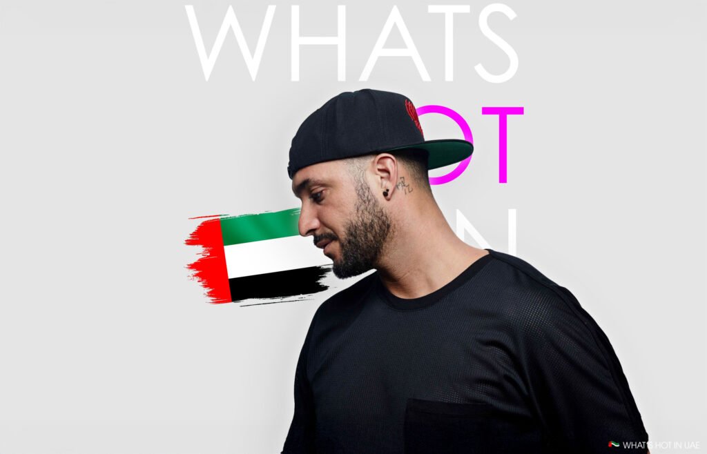 Loco Dice in Dubai wearing a black cap and shirt, standing against a background with a stylized UAE flag and partially visible text.