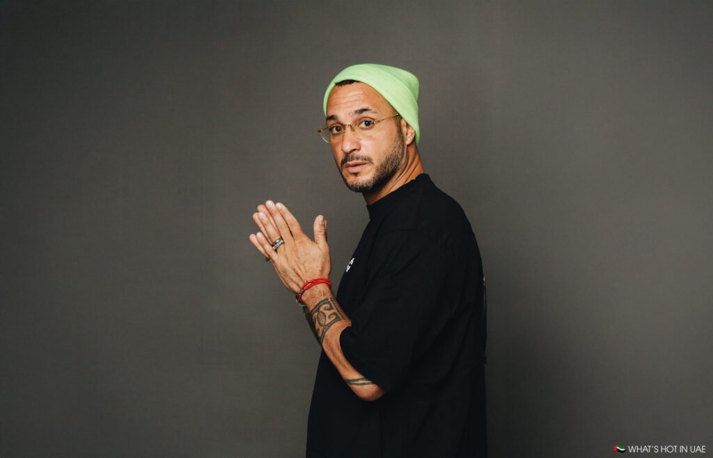 A person in a black shirt and green beanie, wearing glasses and displaying tattoos on both arms, stands against a plain background.