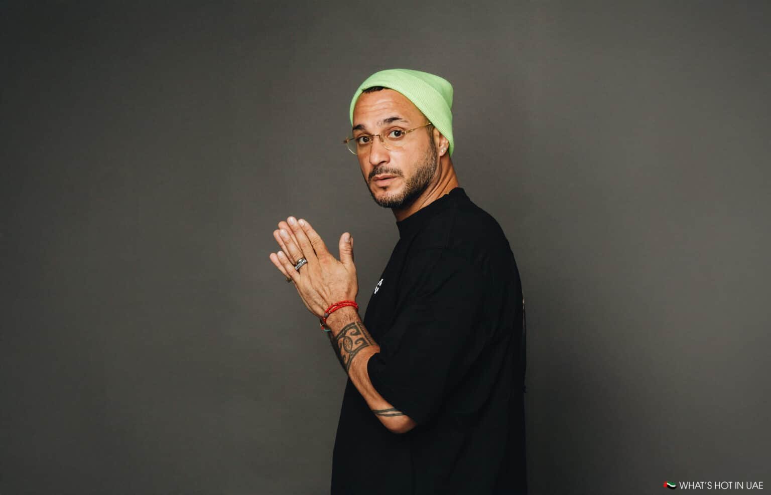 A person in a black shirt and green beanie, wearing glasses and displaying tattoos on both arms, stands against a plain background.