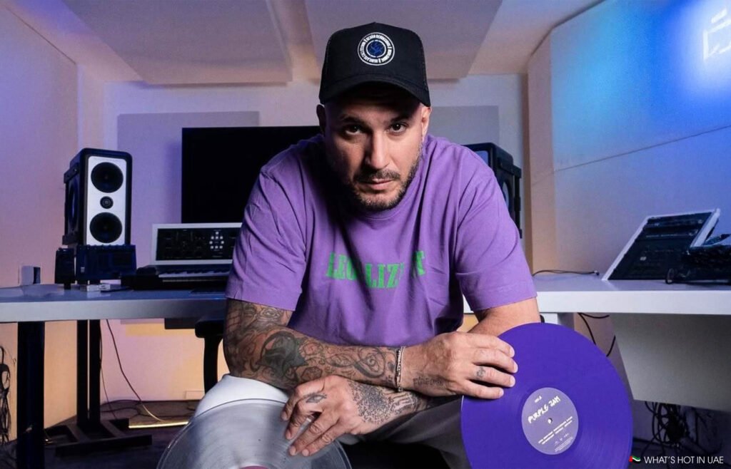 A man wearing a purple shirt and black cap holds vinyl records in a music studio with speakers and monitors.