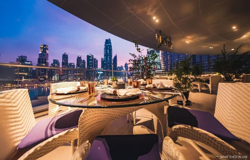 Elegant outdoor dining setup with wicker chairs and a round table, overlooking a city skyline at dusk.