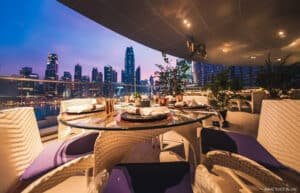 Elegant outdoor dining setup with wicker chairs and a round table, overlooking a city skyline at dusk.