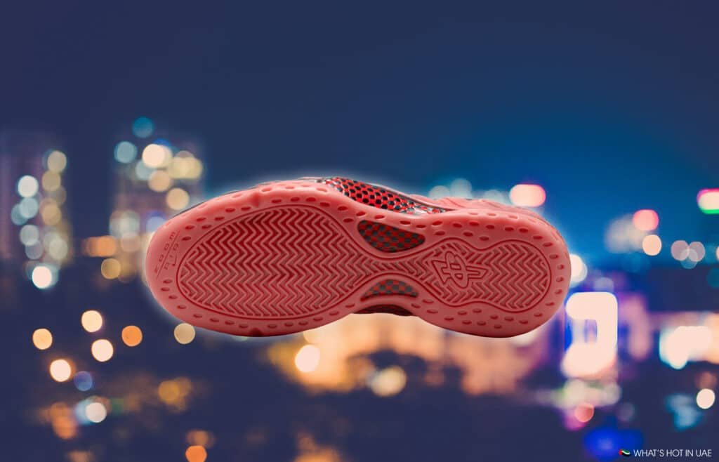 The underside of a pink sneaker sole with a textured pattern is prominently displayed against a blurred cityscape at night.