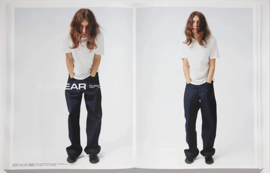A person with long hair wearing a white t-shirt and baggy NO FEAR SPORT Essentials jeans poses against a plain white background.