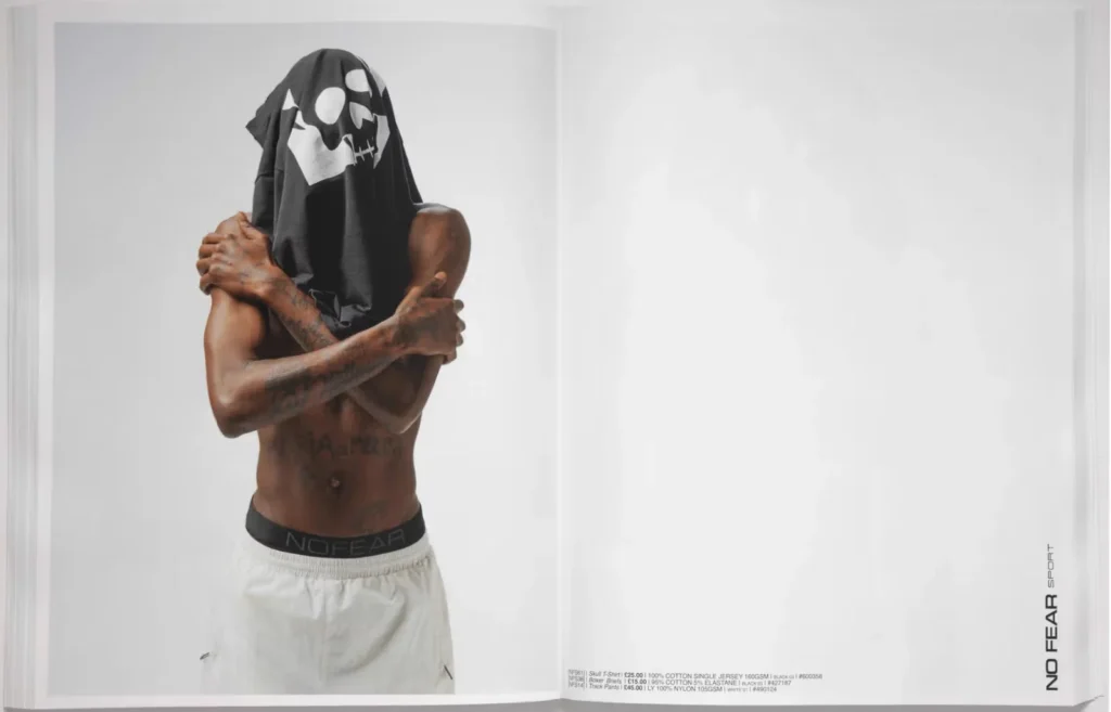 A person with a shirt over their head, wearing white pants and a black waistband with "No Fear" text, stands against a plain background.