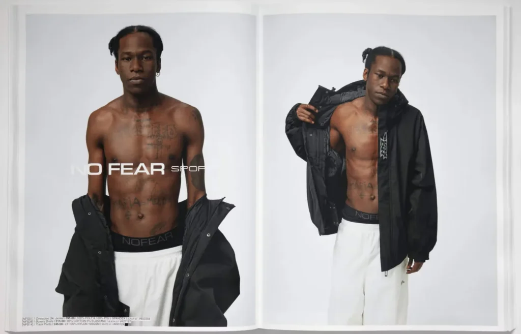 A shirtless man with tattoos poses for a fashion shoot, wearing a black NO FEAR SPORT Essentials jacket and white pants against a plain background.