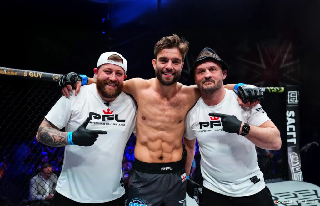 Three men smiling, with the central figure, Alfie Davis shirtless, standing inside a cage marked "Professional Fighters League."