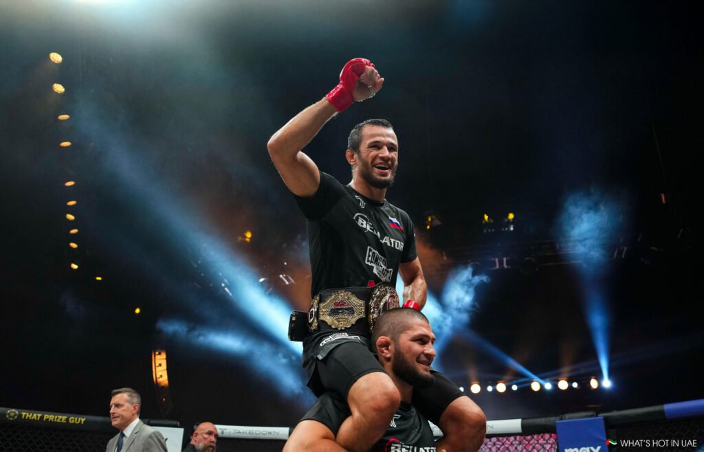 A triumphant Nurmagomedov wearing a championship belt is lifted on another fighter's shoulders in a spotlighted arena. 