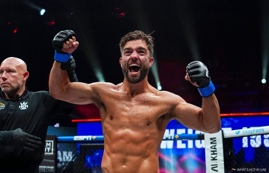 A triumphant mixed martial artist celebrates in the ring with arms raised, wearing black gloves.