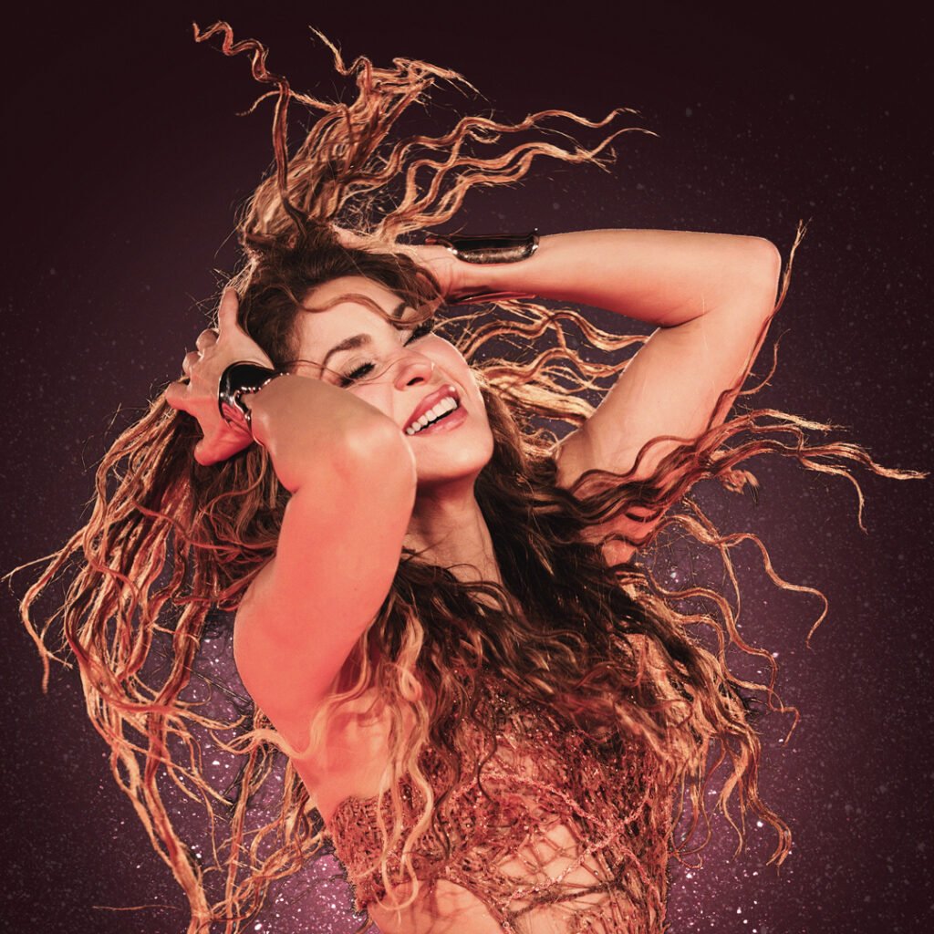 A person with long, wavy hair joyfully posing with hands on the head against a sparkling, reddish-pink background. Global Superstar Shakira OFFLIMITS Music Festival