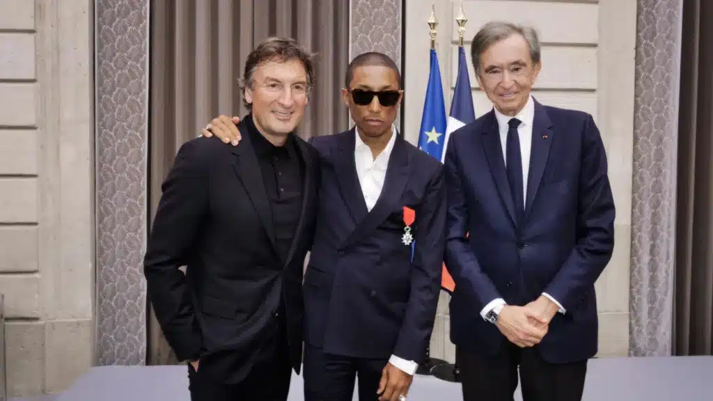 Three men in suits stand together, with the central figure wearing sunglasses and a medal, in an elegant room with flags in the background. Pharrell Williams' Légion d'honneur