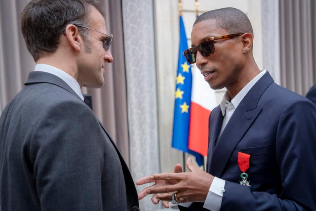 Two men in suits converse in front of French and EU flags, wearing sunglasses and one with a medal on his lapel.