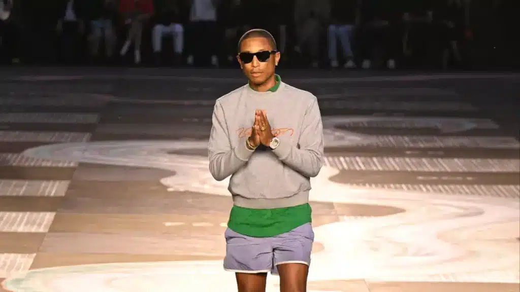 A person in sunglasses walks the runway wearing a gray sweatshirt, green shirt, and purple shorts, clasping their hands together.