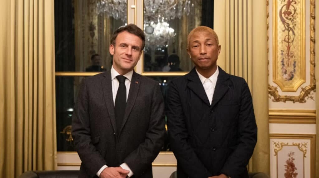 Two men in suits stand side by side in an elegant room with a chandelier and ornate walls.