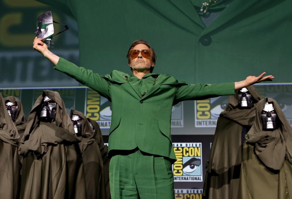 A man in a green suit spreads his arms onstage, surrounded by hooded figures in masks, with Comic-Con signage in the background.