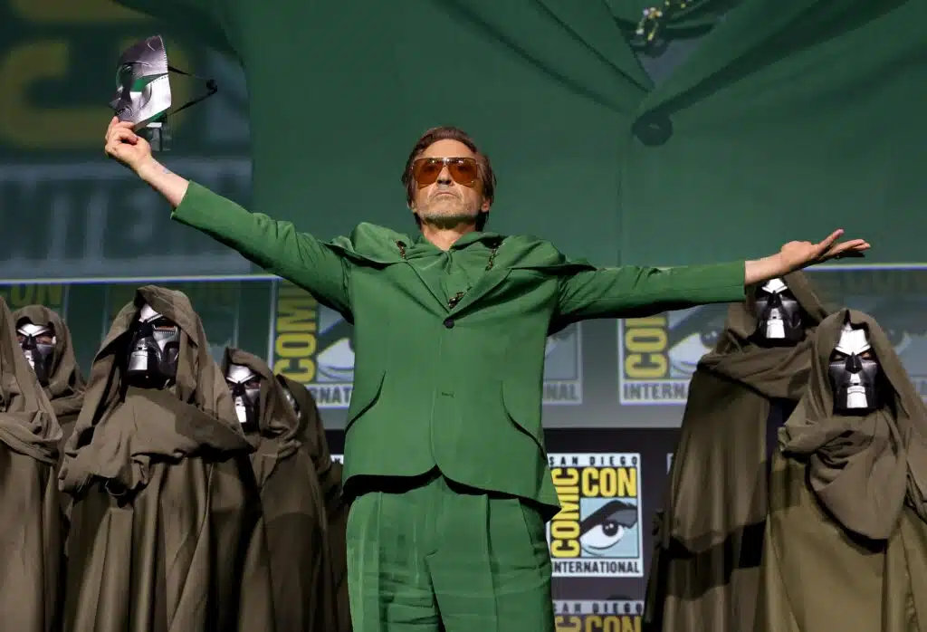 A man in a green suit spreads his arms onstage, surrounded by hooded figures in masks, with Comic-Con signage in the background.