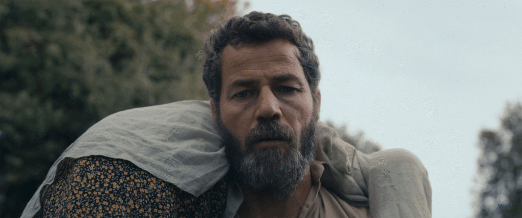 Film still showing a bearded man outdoors carrying a woman over his shoulder, with a natural landscape in the background.