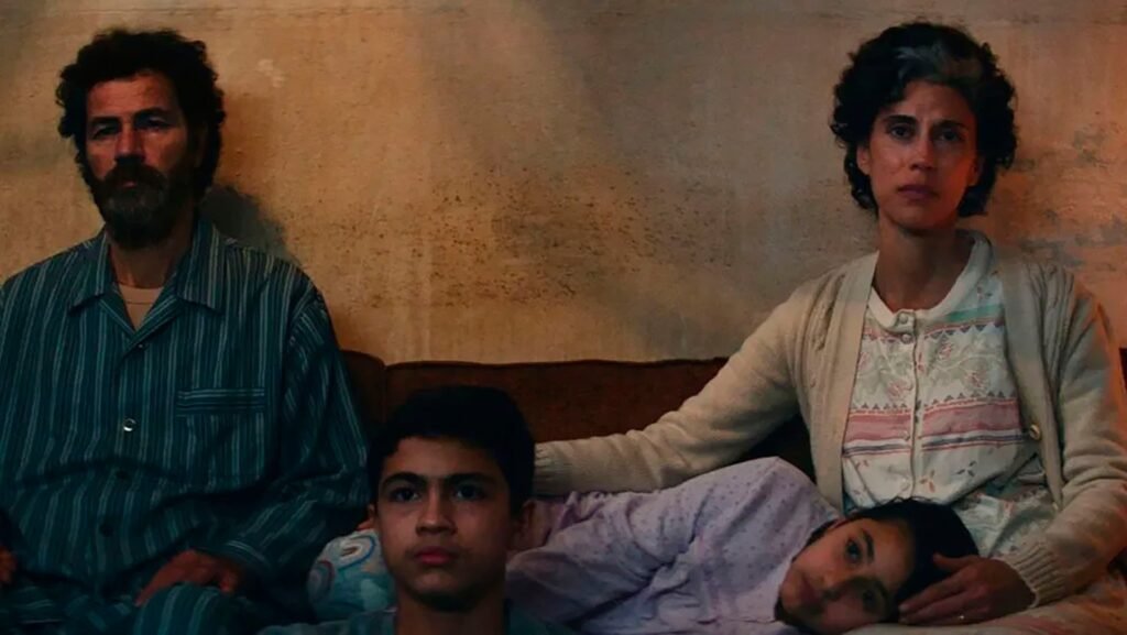 Film still depicting a family seated closely together indoors, with two adults and two children sitting on a sofa in a dimly lit room.