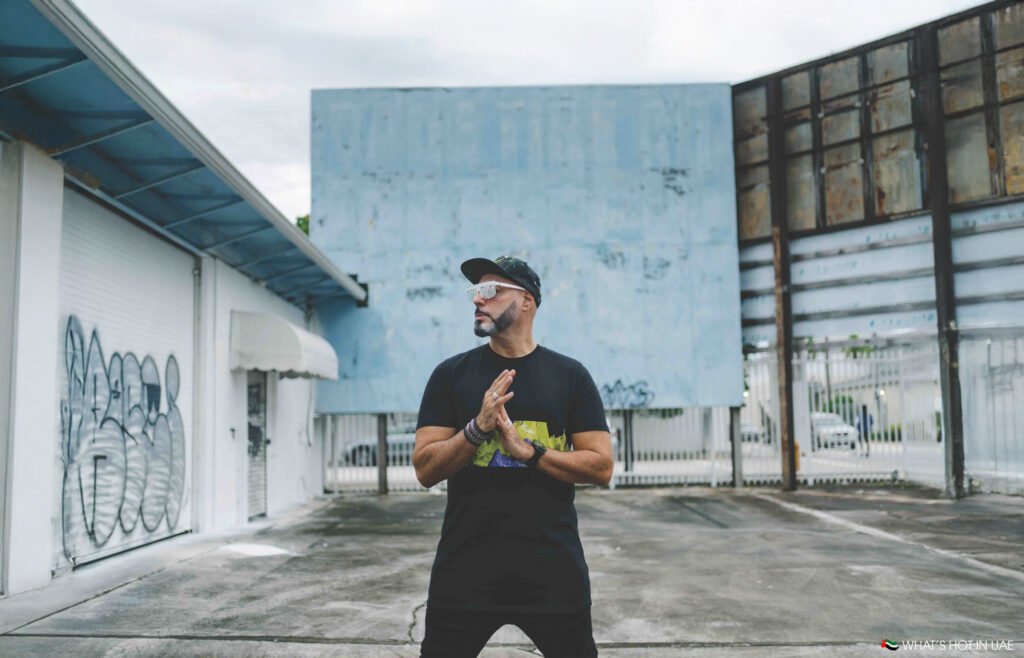 Roger Sanchez wearing a black t-shirt and cap stands on an urban street with graffiti and weathered buildings in the background.