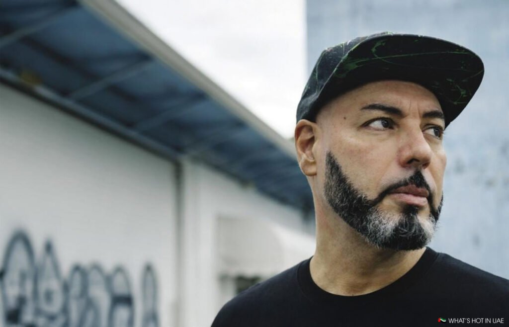 Roger Sanchez with a beard and cap looks to the side, standing outside near a graffiti-covered wall.