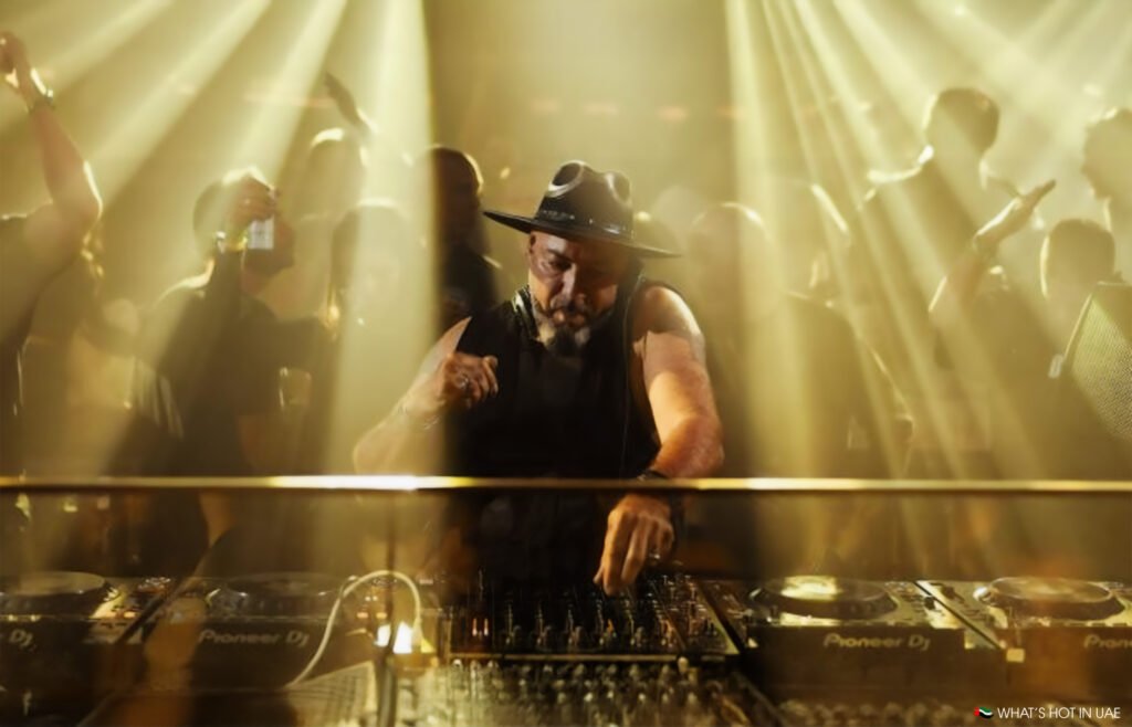 Roger Sanchez performing energetically in a club, surrounded by a crowd with spotlight beams creating a dynamic atmosphere.