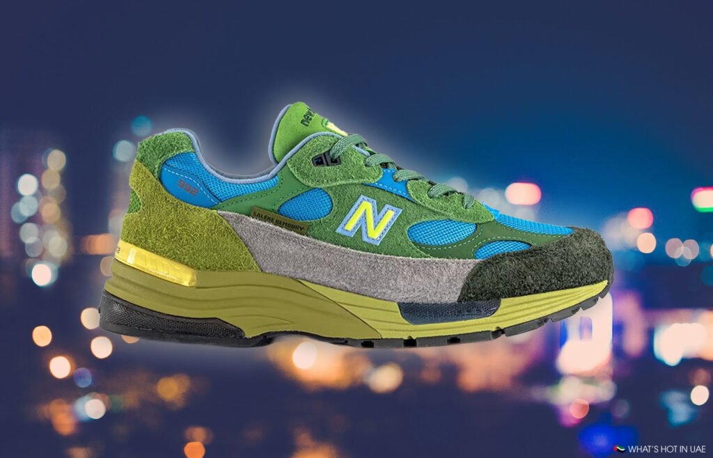 A colorful sneaker with a green, blue, and gray design features the "N" logo, against a blurred urban night backdrop.