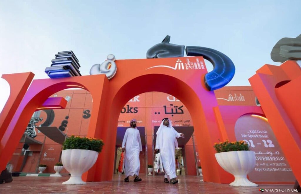 Two people in traditional attire walk through a large, colorful archway adorned with books and Arabic text.